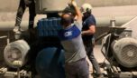 Uzman bir teknisyen, endüstriyel blower sisteminin parçalarını dikkatlice incelediği bir atölyede çalışıyor. Görselde teknisyenin elinde tuttuğu blower pervanesi ve yanında açık motor kısımları görülebilir, bu da periyodik bakımın detaylı bir süreç olduğunu vurgular.