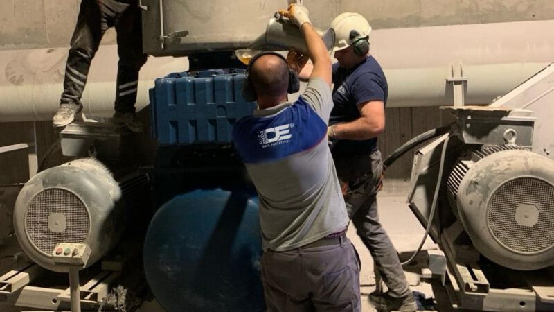 Uzman bir teknisyen, endüstriyel blower sisteminin parçalarını dikkatlice incelediği bir atölyede çalışıyor. Görselde teknisyenin elinde tuttuğu blower pervanesi ve yanında açık motor kısımları görülebilir, bu da periyodik bakımın detaylı bir süreç olduğunu vurgular.