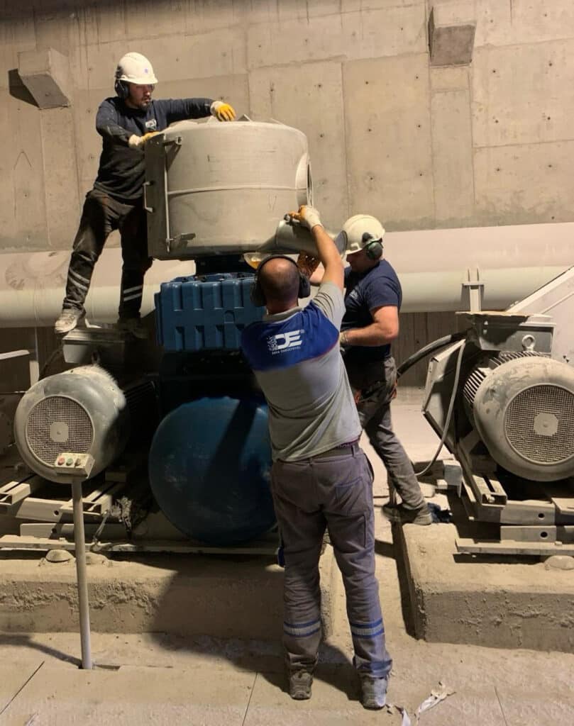 Uzman bir teknisyen, endüstriyel blower sisteminin parçalarını dikkatlice incelediği bir atölyede çalışıyor. Görselde teknisyenin elinde tuttuğu blower pervanesi ve yanında açık motor kısımları görülebilir, bu da periyodik bakımın detaylı bir süreç olduğunu vurgular.